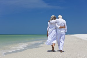 A senior couple with their arms wrapped around one another walks along a sandy tropical beach. They are both wearing all white, with the exception of their footwear. In the background, a clear blue sky provides contrast to the all-white attire. The tide flows on the shore, nearly touching the couple's feet.